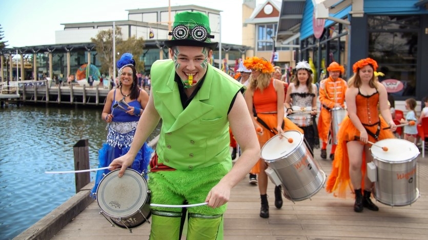 A man in a green vest playing a drum leads a parade of brightly dressed people with instruments along a waterside location