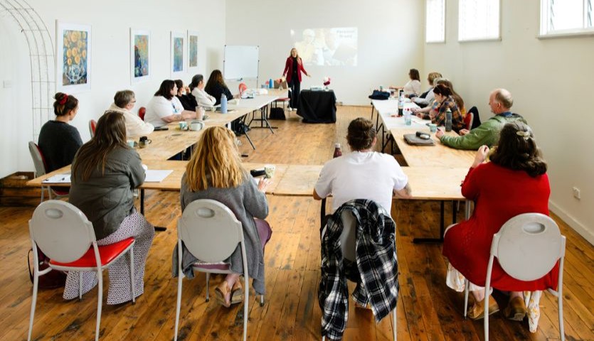 A group of creatives sit at desks, watching a woman presenting with a whiteboard.