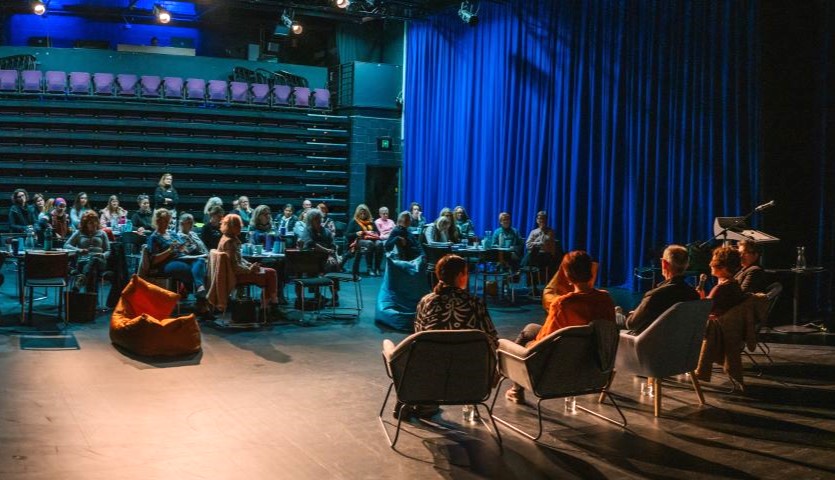 Four people sit on chairs in an auditorium with their back to the camera, presenting to a group of people who are sitting at tables. 