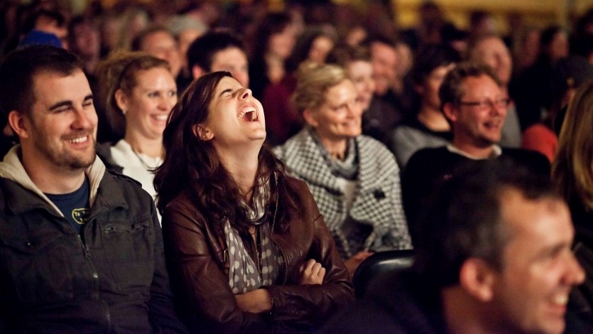 A crowd of people sitting in an auditorium are all laughing at something they can see out of frame