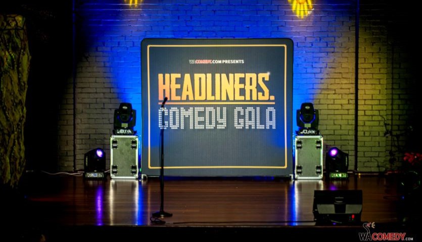 A sign that says "Headliners Comedy Gala" stands on a stage, framed by yellow and blue lights and with sound equipment on either side