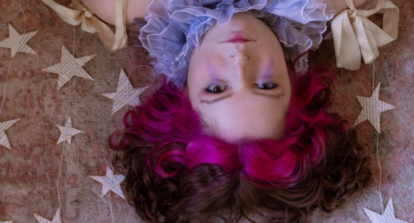 a young person with pink and black hair lies on their back with paper stars cut out on the ground beside them