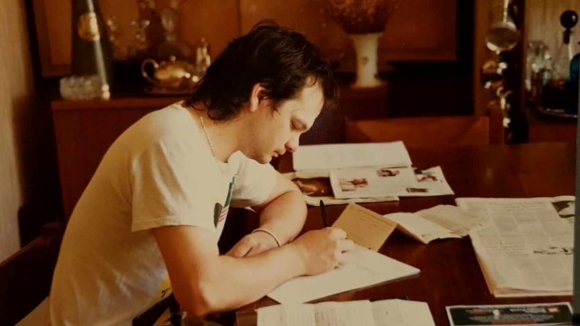 A young man in a white shirt leans over a desk, writing music notation onto to paper