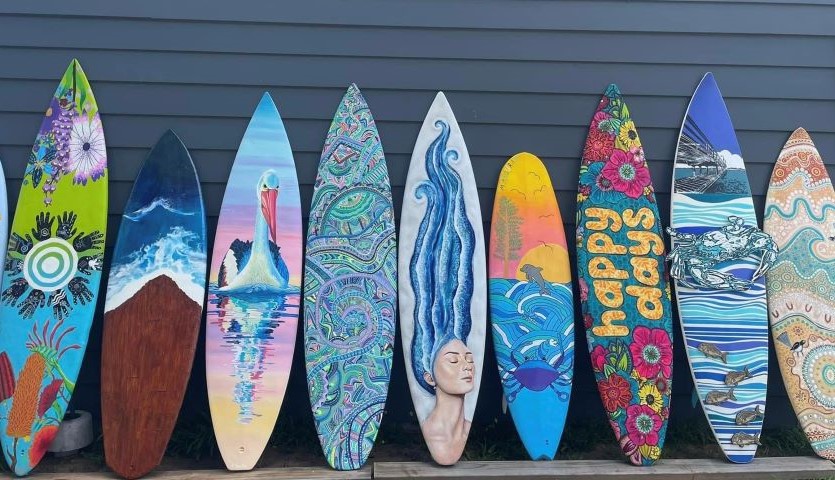 A row of 9 painted surfboards, depicting different water-based pictures, lean up against a grey wall