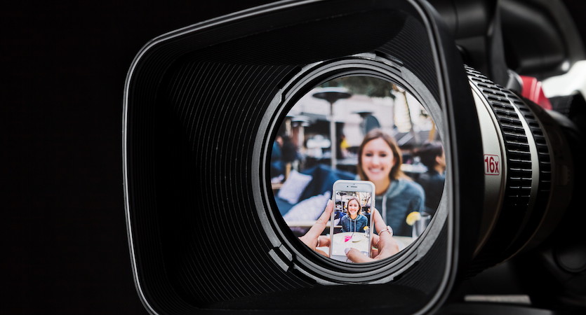 A close up view of the lens of a video camera. Reflected in the lens is a scene of a woman at a cafe, being filmed on a phone camera