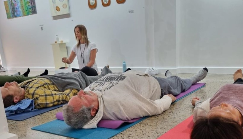 A group of people lie on Yoga-mats on the floor of an art gallery, with one person sitting at the front leading a meditation