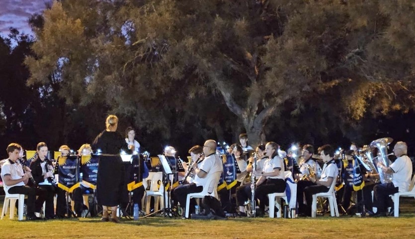 A large concert band wearing white polo shirts sit outdoors on a grassed area playing their instruments