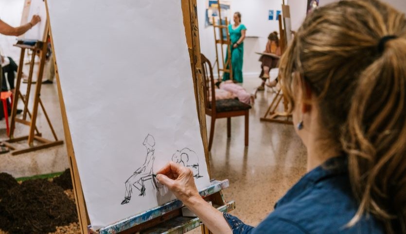 The back of one woman's head in the foreground, as she sketches a human form on a piece of paper on an easel. In the background are other people drawing.