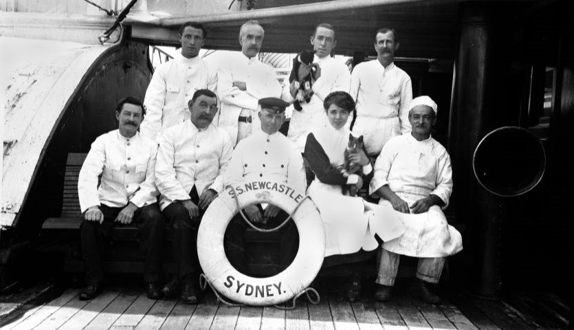an old fashioned, black and white photograph of people in uniform on the deck of a ship, with a life-saver that says "SS Newcastle" and a dog held in one man's arms