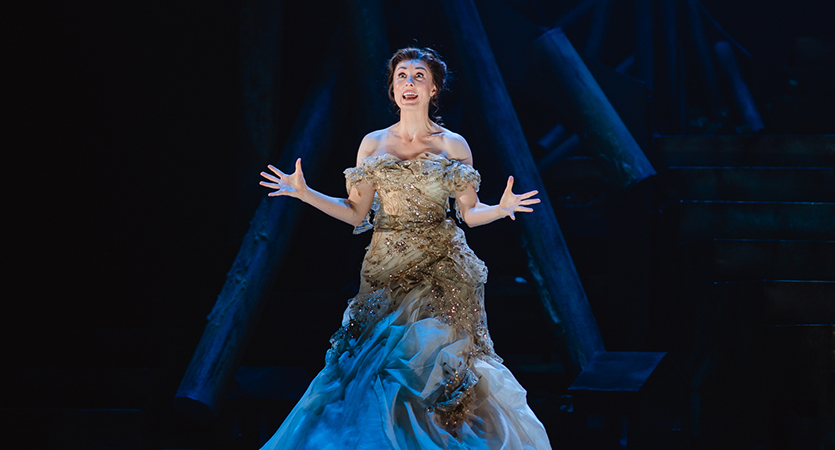 A woman stands onstage in a gold and blue dress, holding her arms out to the side with a passionate expression on her face as she sings