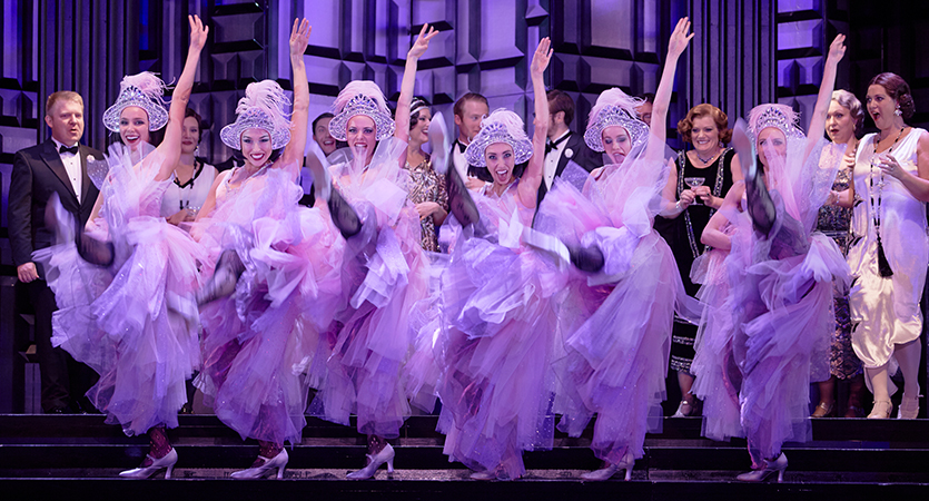 A line of dancers wearing white, feathered dresses are all onstage, kicking their feet in the air in unison with happy smiles on their faces