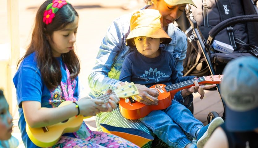 Two small children, around 2 and 6 years old, sit with their mother, holding and playing ukeleles at an outdoor location