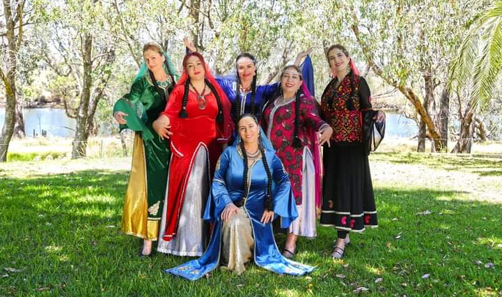 6 dancers wearing long colourful dresses with big sleeves are posed in an outdoor, grassed location