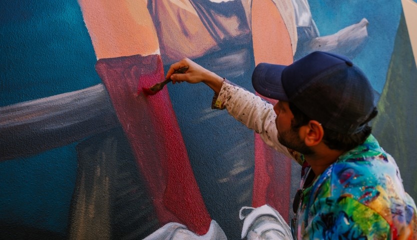 A man wearing a baseball cap and a colourful shirt is painting a mural of a young boy wearing red socks, in an outdoor location