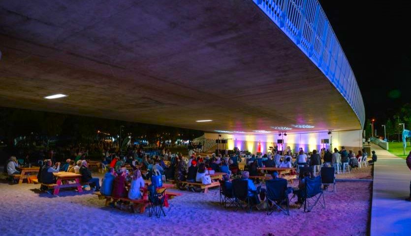 A large audience sit beneath a bridge on picnic chairs at night time, watching a performance of an opera singer