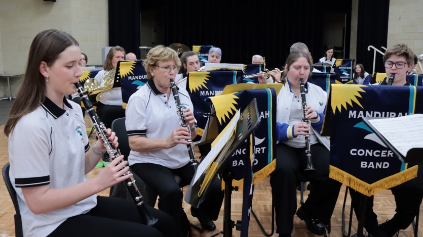 A group of people playing wind instruments are seated in a large hall, looking at music on their music stands