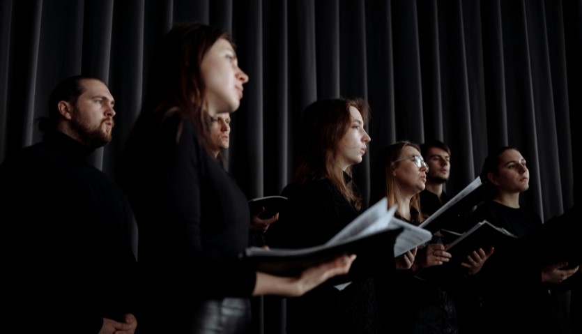6 people wearing black, standing against a black curtained background, are holding songbooks and singing