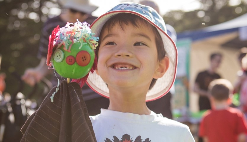 A young boy wearing a bucket hat stands outside with a big smile on his face showing missing front teeth. He is holding a hand puppet made from a green apple