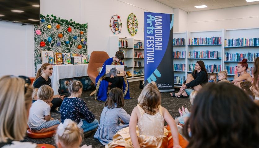 A group of children sit on a carpet in a library, looking at a young woman wearing a blue cape, who is reading a story to them.