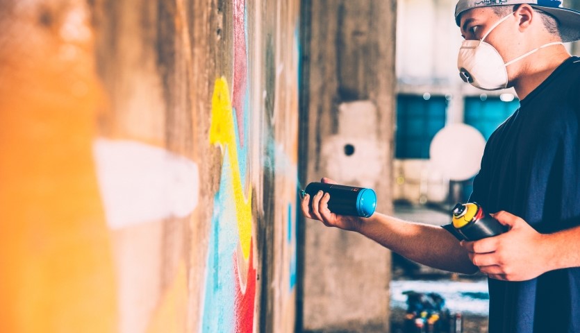 A graffiti artist wears a respiration face mask as he spray paints a colourful design onto a concrete wall