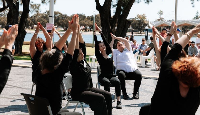 A group of people wearing black and white are seated outside in a spiral formation, with their arms held above their head in a dance move