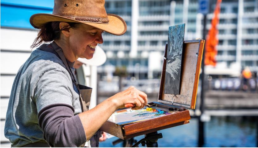 a smiling woman wearing an akubra paints on a small board in an outdoor, waterside location