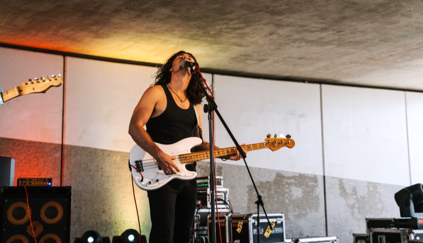 A man with long hair is playing electric guitar on a stage surrounded by sound equipment. The background is a concrete wall and low ceiling.