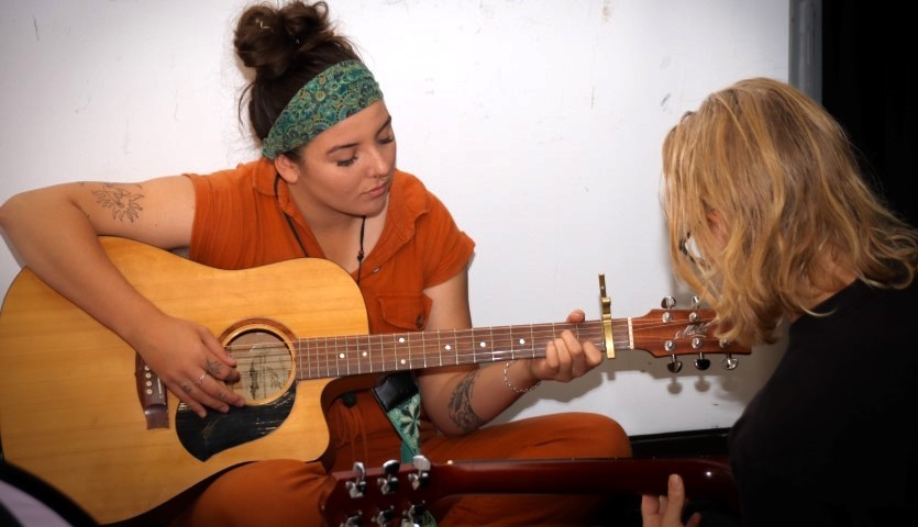 A young woman in an orange shirt is sitting, playing a guitar. In front of her is the back of a woman's head, who is looking at what she is playing as if giving feedback. 