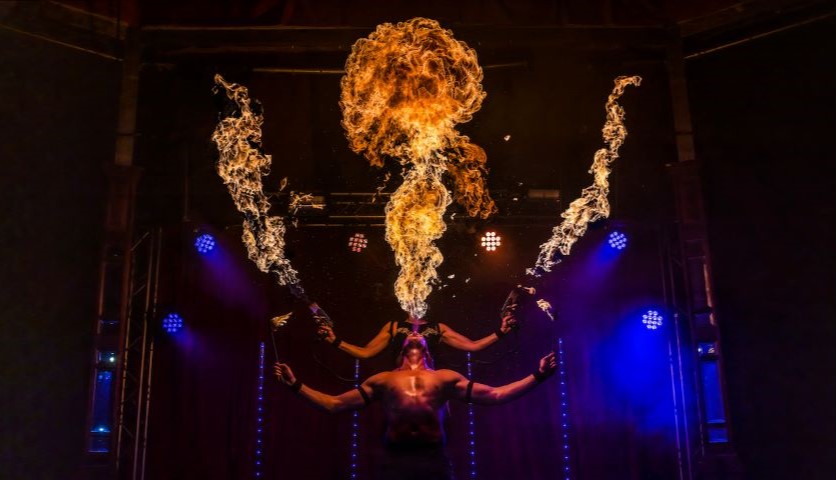 Two circus performers breathe fire and hold fire sticks in their hands, on a dark stage with dramatic blue and red lighting