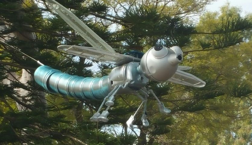 A blue and silver sculpture of a dragonfly, seemingly made from scrap metal, hangs suspended in a tree.