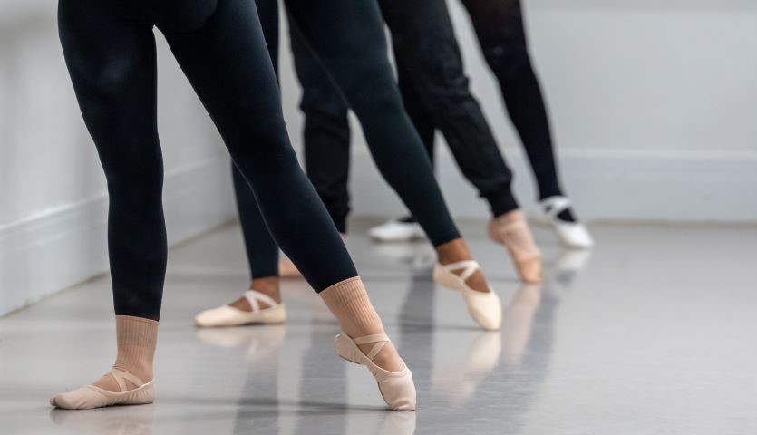 Four sets of legs wearing black tights and ballet shoes are lined up, all are pointing their left toes to the side on the ground.