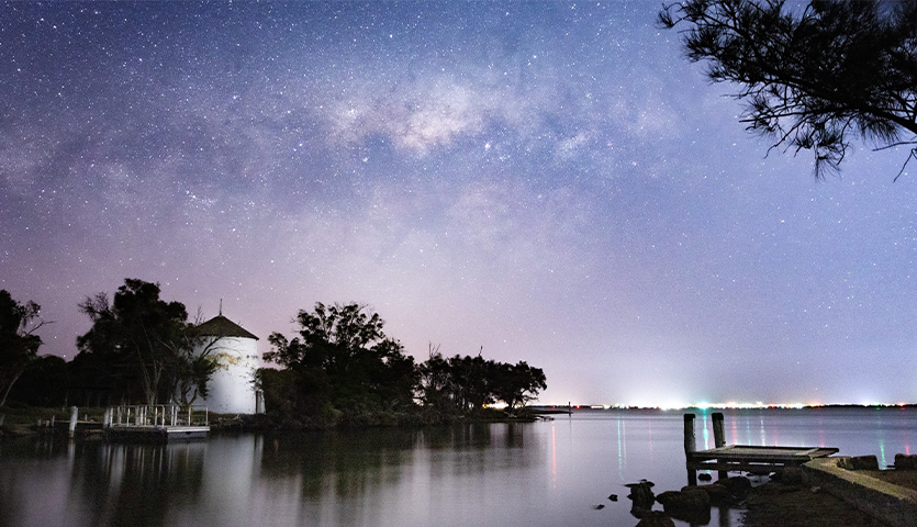 The night sky glistens with stars over a body of water, which reflects the sky's blue and purple hues