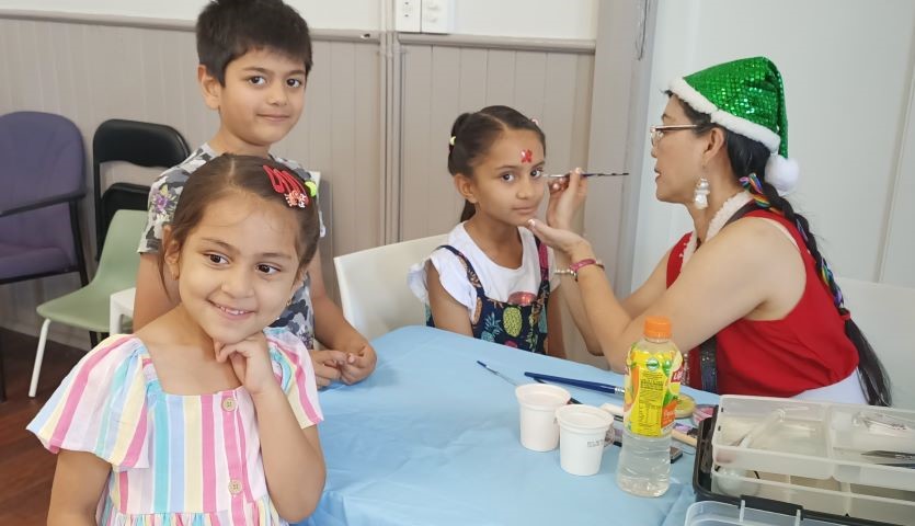 Three children wait in line for a woman in a green, sparkly hat to paint their face. The youngest child, a girl in a pink and blue striped dress, smiles with wonder just past the camera.