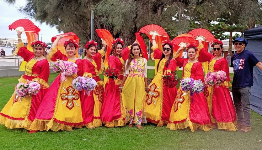 8 women in bright red and yellow dresses, and one man in a black shirt, stand in a line smiling at the camera in an outdoor location. The women are posed with large, ornamental fans.