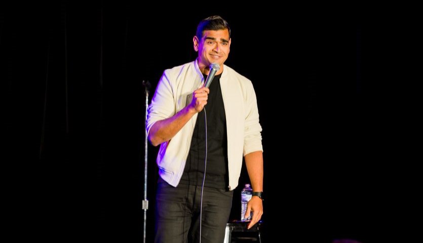 A man wearing a black t-shirt and white un-buttoned shirt stands on a dark stage, smiling wryly and holding a microphone.