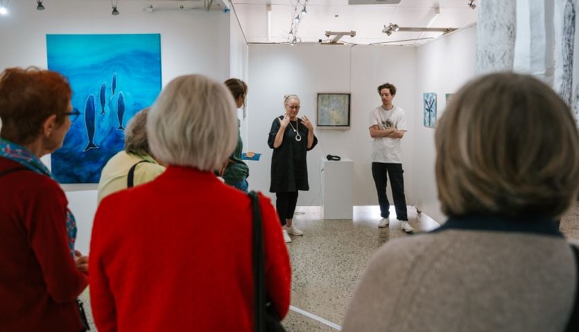 Two artists , a man and woman, stand before a group of people, speaking about the artwork on the wall behind them.