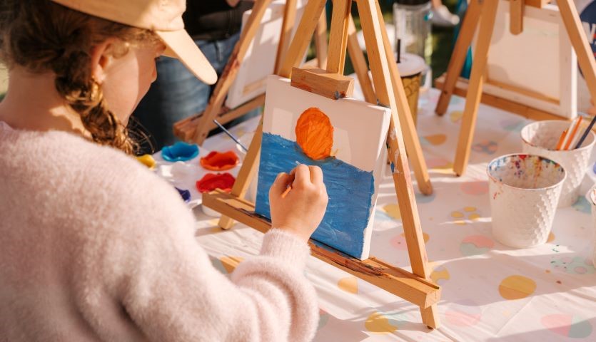 A young girl in a pink jumper and pigtails, paints a picture with blue water and an orange sun.