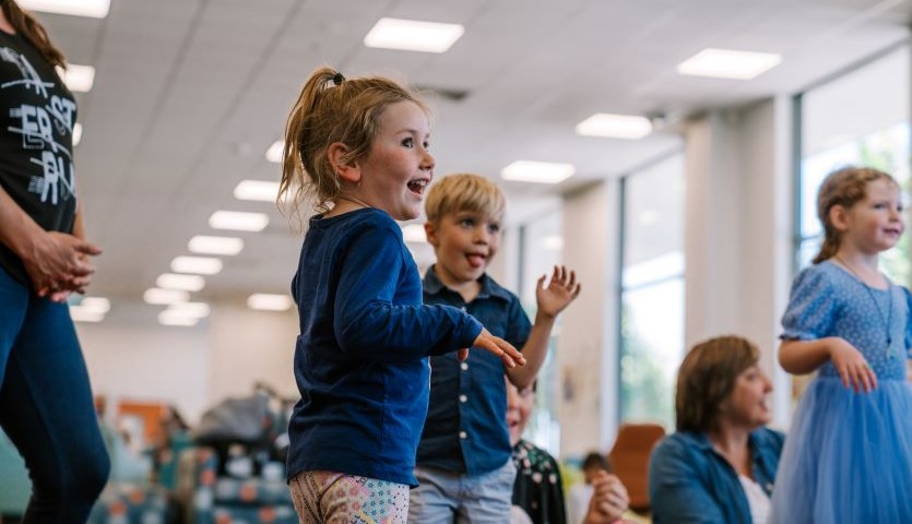 A number of children dance around a library space. The closest child, a small girl, seems to be flapping her hands and poking her tongue out slightly.