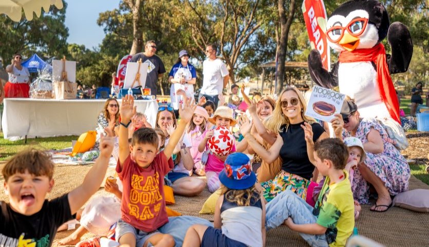 A group of children sit on the ground in a park, with their hands thrown up in celebration. There is also a large penguin mascot and an adult reading to them.