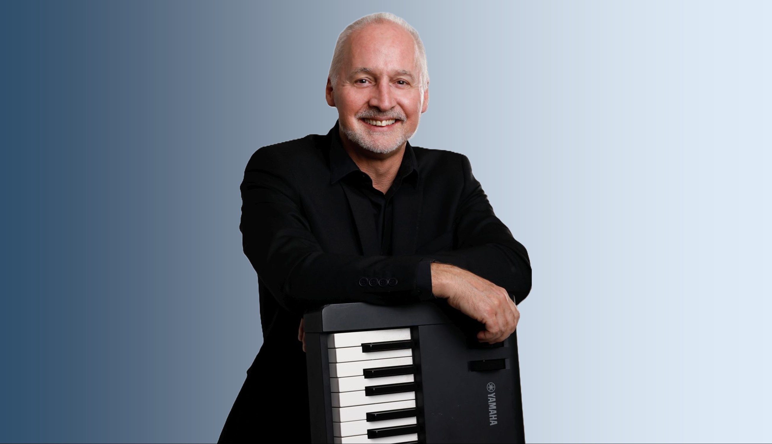 A man leans on a keyboard, which is standing on end. He is smiling, and the background fades from blue on one side to white on the other.