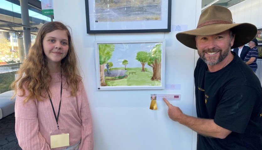 A young girl and a man wearing an akubra stand next to a painting hung on a wall. They are both smiling and the man is pointing to a sign on the wall, indicating that the girl's painting has won a prize.