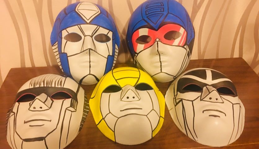 Five masks sit on a wooden table, each are drawn on with thick, black angular lines and bright colours to look like characters from the cartoon series Transformers