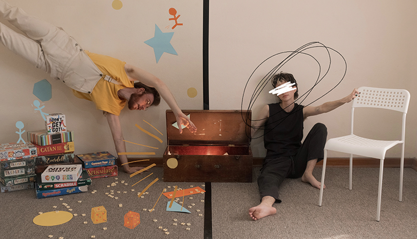 on one half, a man wearing a yellow shirt and overalls is doing a handstand, surrounded by toys. On the other side, a man wearing all black is reclined lazily, and has scribbles covering his eyes an circling his head.