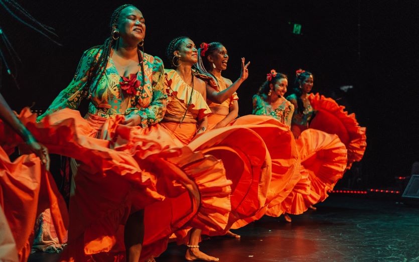 Five women wearing bright orange skirts, colourful shirts and long braids and curls in their hair, are dancing barefoot onstage, swishing their skirts around dynamically and smiling.