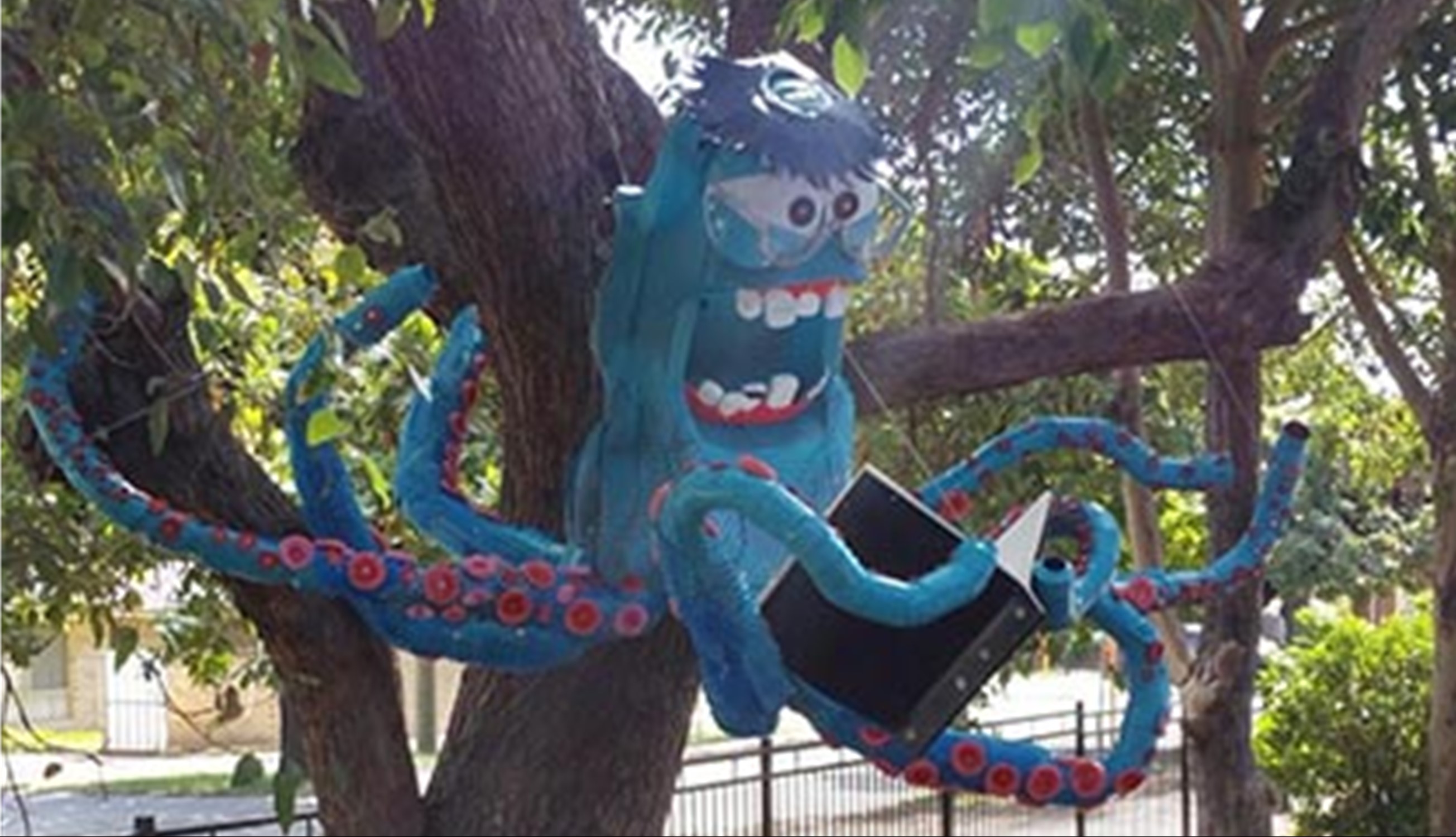 A sculpture of a blue octopus with a wide, manic grin is suspended in a tree