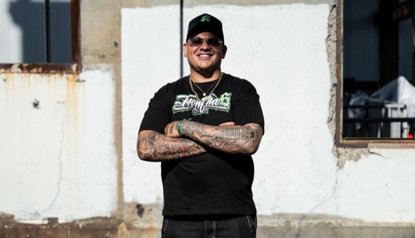 A man in a black t-shirt and black cap stands in a run down urban environment, hands crossed over his chest and smiling widely.