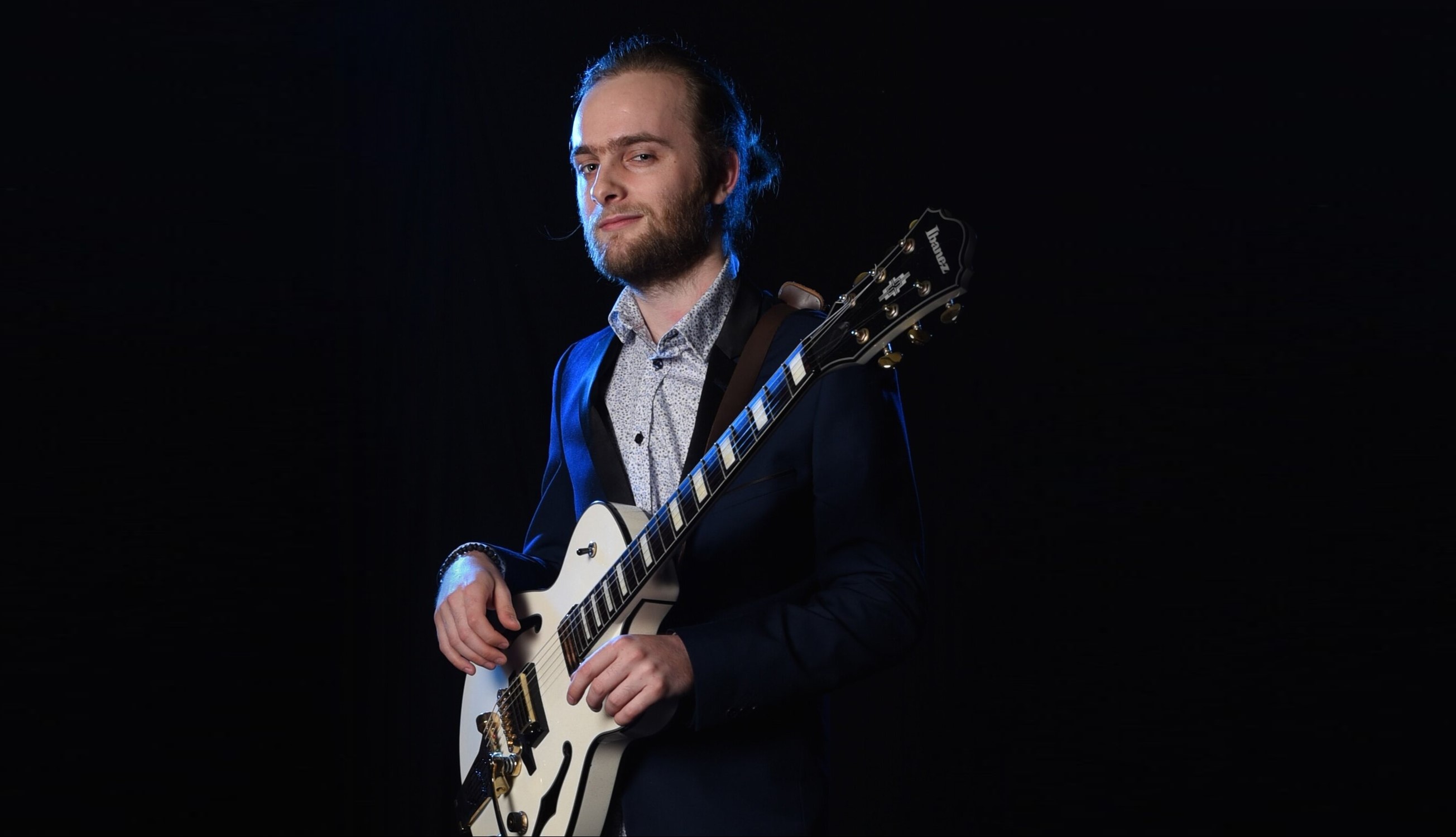 A young, bearded man with a wry smile stands against a black background, wearing a suit jacket and holding an electric guitar