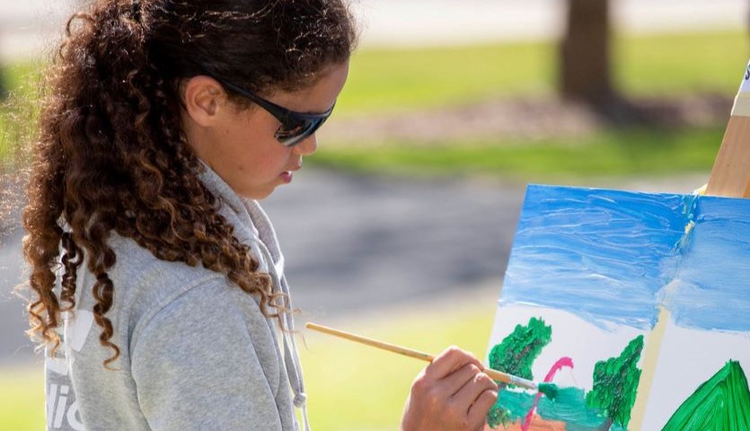 A girl with long, dark curly hair, wearing sunglasses, stands at an easel in an outdoor location, painting a landscape image on her canvas.