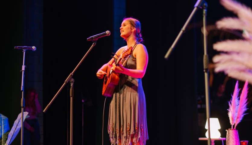 A teenage girl in a brown dress holds a guitar and sings into a microphone, in an onstage setting with soft lighting.