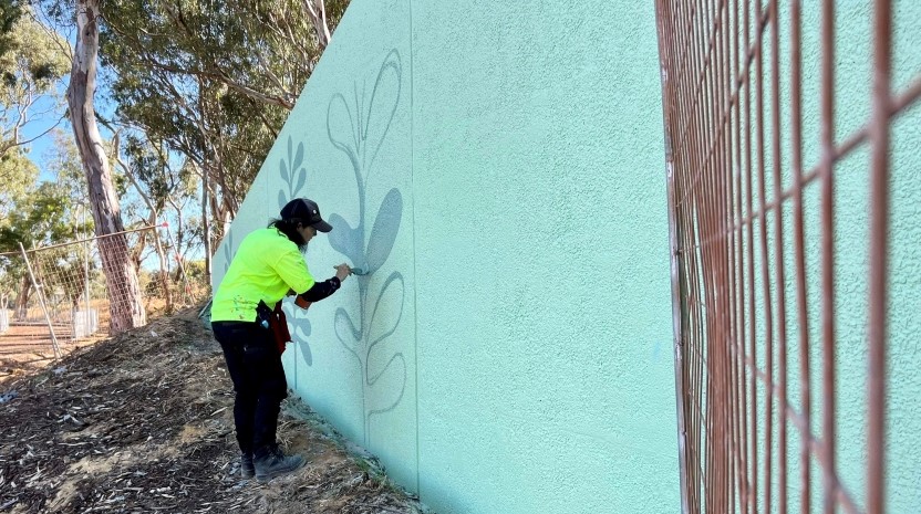 A woman wearing a high-visibility shirt is painting a mural on a green wall in a rural setting